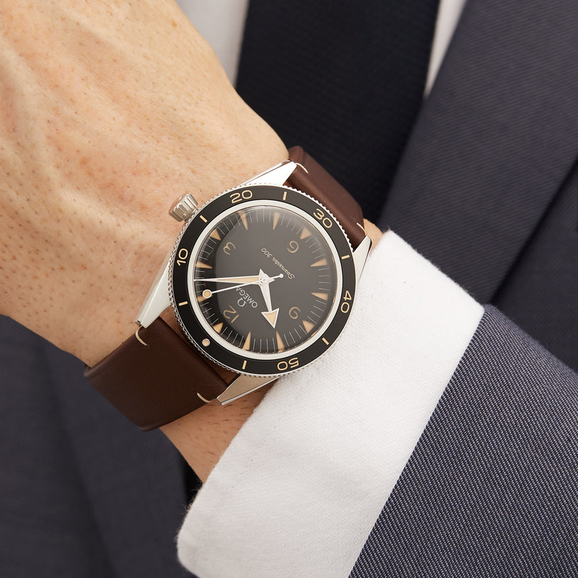 A person wearing an OMEGA Seamaster 300 with a brown leather strap, black dial, and silver case, paired with a white shirt and dark suit jacket.