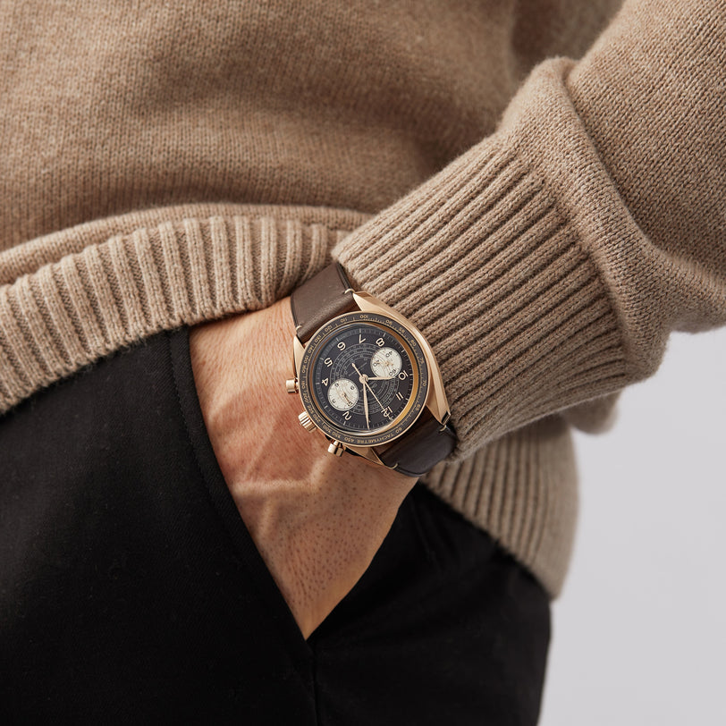 A person in a beige sweater and black pants puts their hand in their pocket, displaying an OMEGA Speedmaster Chronoscope with a gold case, dark dial, and leather strap that highlights its vintage-inspired design.