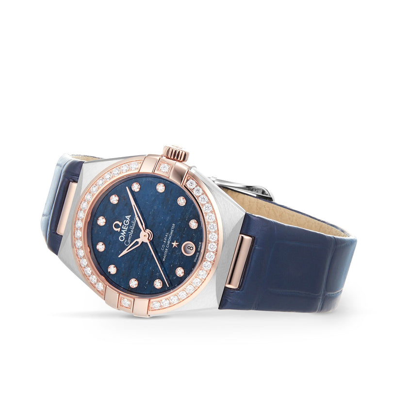 The OMEGA Constellation is a luxury watch with a blue leather strap, diamond-set bezel, blue dial with diamond hour markers, date window, and radiant 18K Sedna™ gold accents.