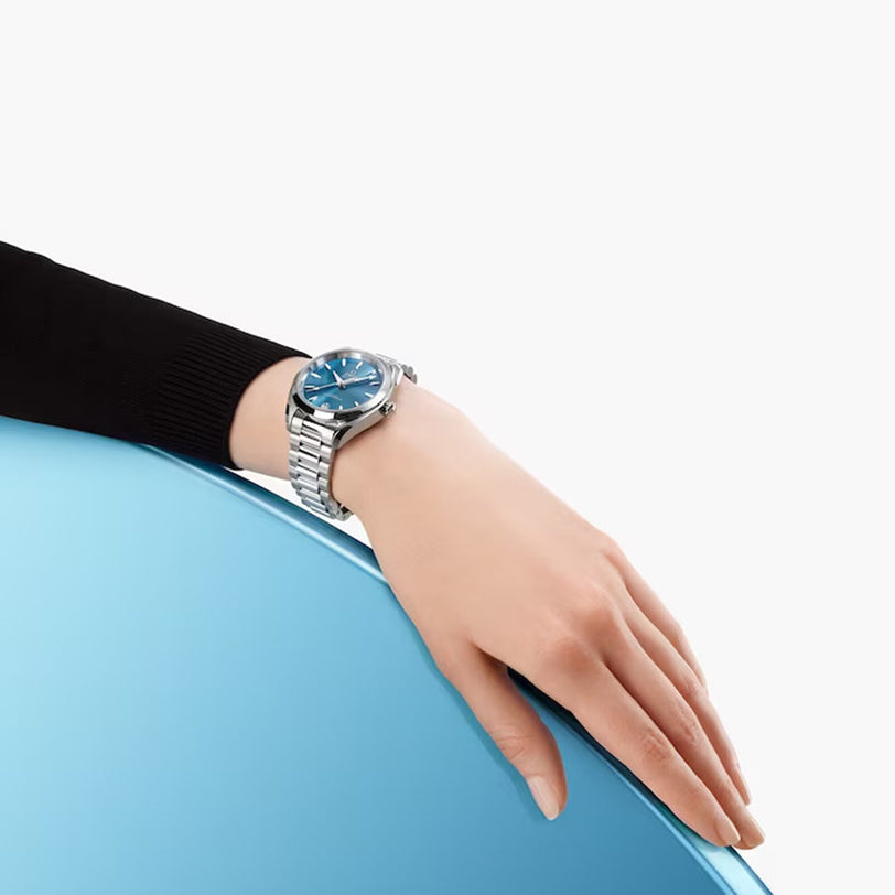 A hand wearing an OMEGA Seamaster Aqua Terra Shades with a blue face rests on a blue surface against a plain white background.