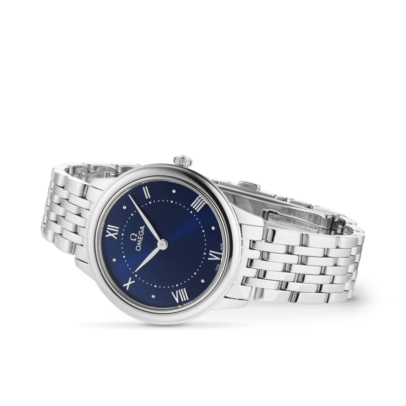 An OMEGA De Ville Prestige wristwatch with a blue dial, Roman numerals, and a stainless steel case with metal link bracelet is shown at an angle on a white background.