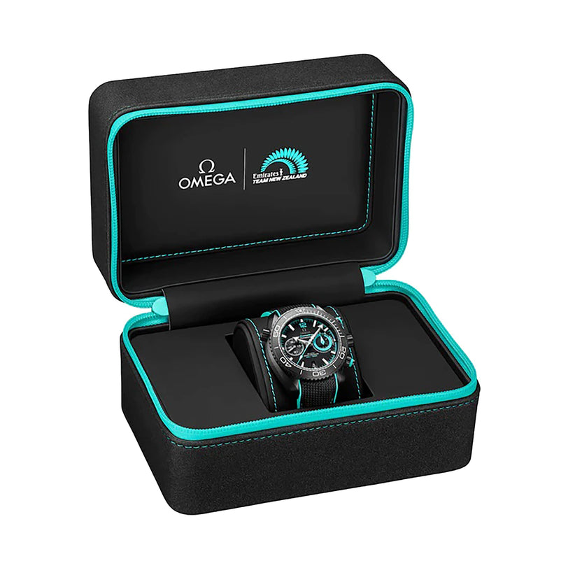 An OMEGA Seamaster Planet Ocean 600M wristwatch with teal accents is displayed in an open black case with teal trim, featuring OMEGA and Emirates Team New Zealand logos inside the lid. Powered by the OMEGA 9900 calibre.