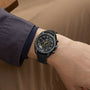 A person wearing an OMEGA Speedmaster Moonwatch Apollo 8 Dark Side Of The Moon with a dark strap, dressed in a gray jacket and tan pants, rests their arm on a surface.