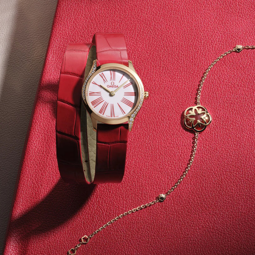 An OMEGA De Ville Mini Trésor wristwatch in 18K Moonshine™ Gold with a white face, red Roman numerals, and a red leather strap is shown beside a gold floral bracelet on a red textured surface. Powered by calibre 4061.