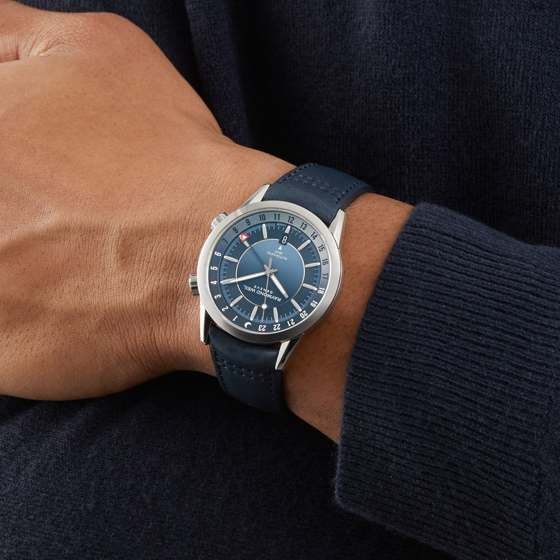 A person wearing a Raymond Weil Freelancer GMT Blue Dial wristwatch with a leather strap, dressed in a dark sweater.