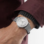 A person wearing a Raymond Weil Millesime Automatic Small Seconds Silver Sector Dial watch with a grey leather strap and white dial, dressed in a maroon jacket and dark sweater.