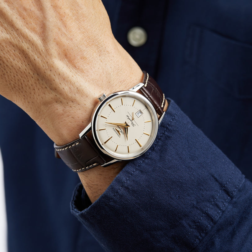 A person in a blue shirt wearing a Longines Flagship Heritage watch with a stainless steel case, brown leather strap, and cream-colored dial.