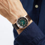 A person in a dark blue shirt showcases the Longines Legend Diver Bronze, featuring a green-black dial with Super-LumiNova markers, a silver bezel, and a dark brown leather strap.