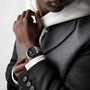 A person in a grey suit and white hoodie wears a Longines Spirit Zulu Time watch with a stainless steel case and black face on their left wrist.