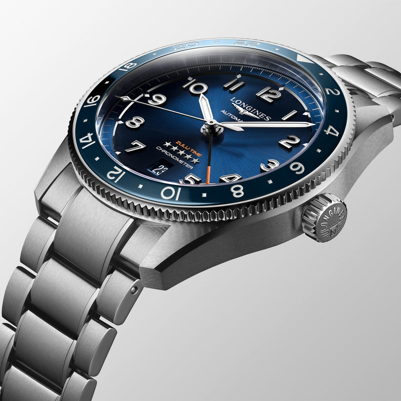 The Longines Spirit Zulu Time showcases a stainless steel case and bracelet, blue dial with Swiss Super-LumiNova hands, Arabic numerals, date window, and a blue bezel—all set against a light background.