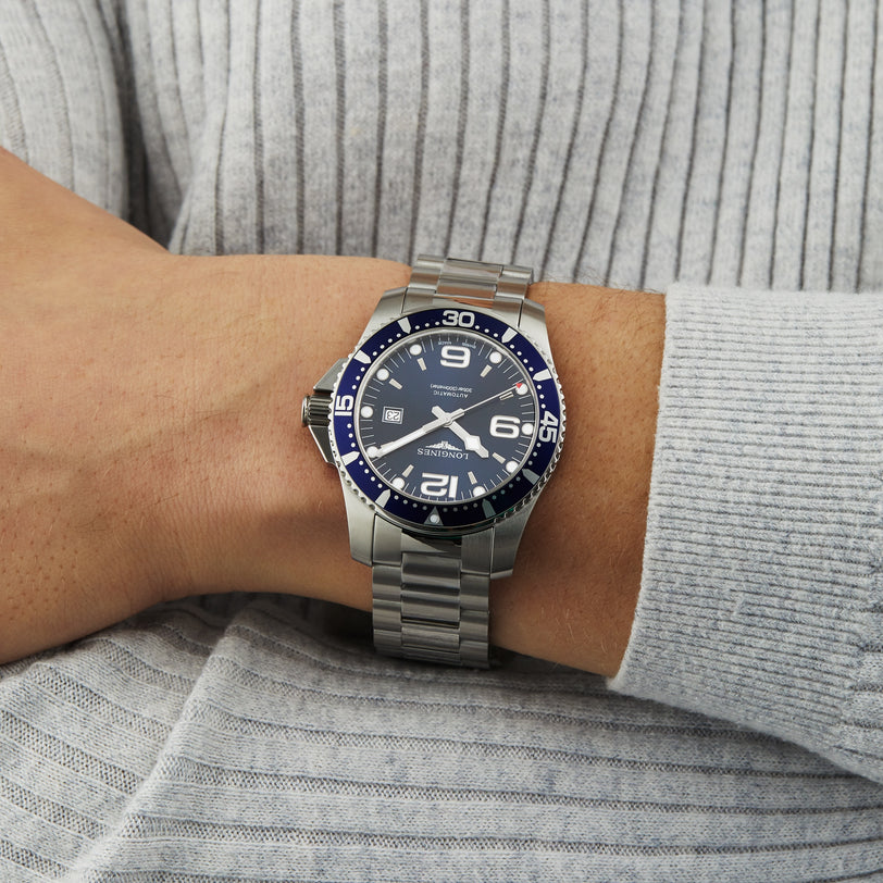 A person wearing a Longines HydroConquest silver wristwatch with a blue face and metal band, powered by automatic movement, rests their arm on a light gray ribbed fabric garment.