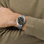 A person in a brown sweater shows off a Longines Spirit Zulu Time watch with a black dial, yellow numerals, green ceramic bezel, and stainless steel case on their left wrist.