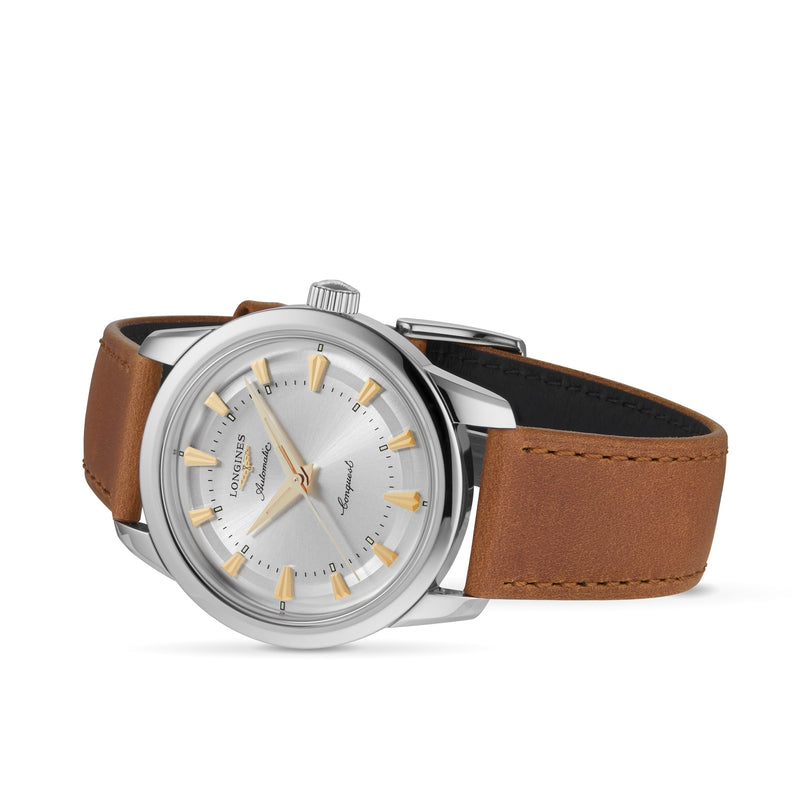 A Longines Conquest Heritage watch with silver dial, gold markers, brown leather strap, and stainless steel case is angled on a white background, showcasing the brands renowned precision.