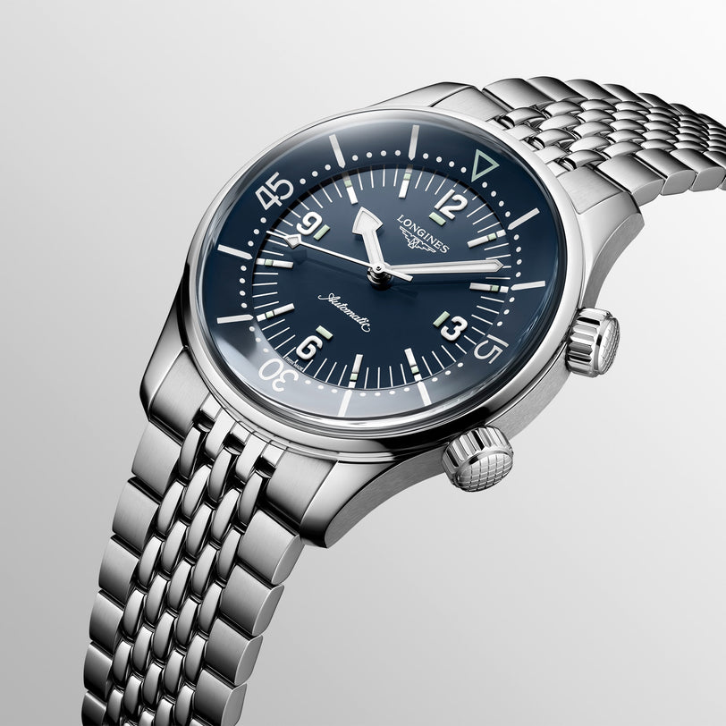 The Longines Legend Diver features a stainless steel case, blue dial with white numerals, two crowns, and a woven metal bracelet—an elegant vintage-inspired automatic dive watch set against a light gradient backdrop.