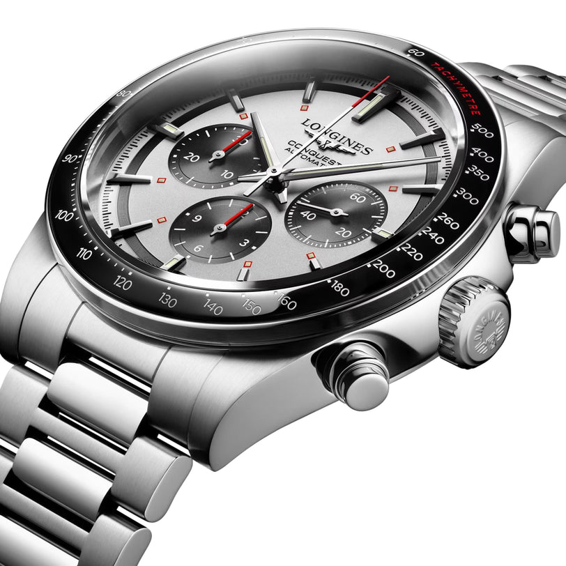 The Longines Conquest Chronograph features a stainless steel case, silver dial with three subdials, red accents, tachymeter scale, metal bracelet, and a precision self-winding movement.