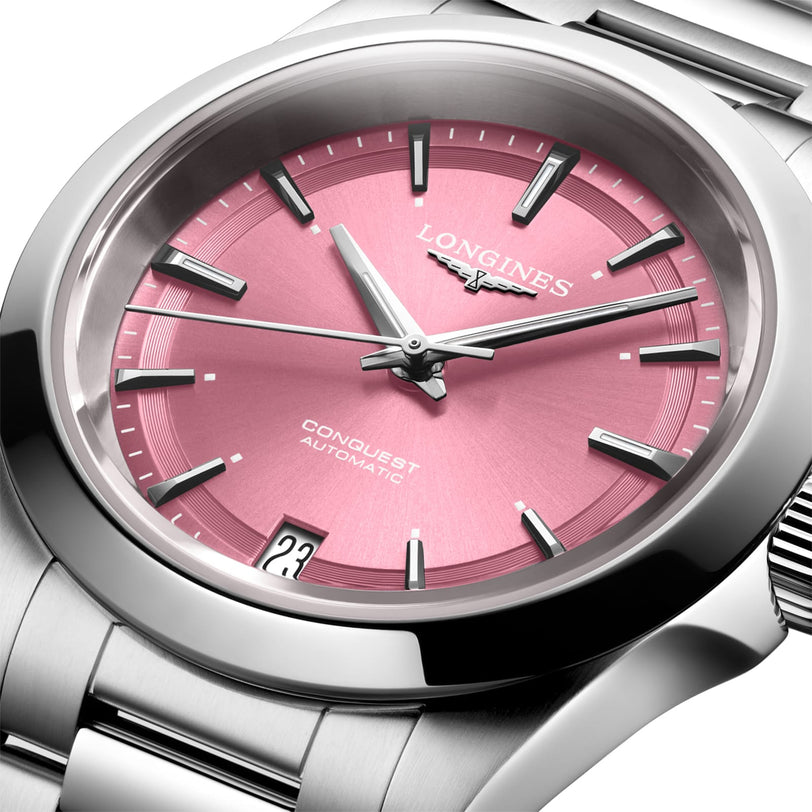 A close-up of the Longines Conquest watch featuring a stainless steel case, pink dial, automatic movement, and a date display at 6 o’clock.