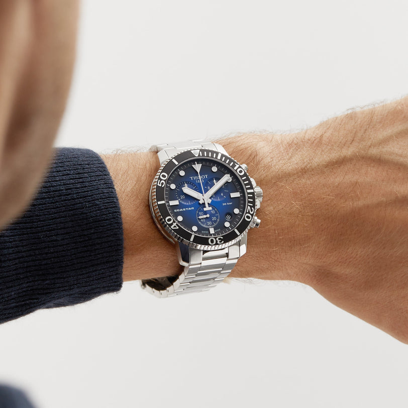 A person wears a Tissot Seastar 1000 Chronograph with a silver case, blue dial, and black bezel on their left wrist.