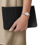 A person wearing a Tissot Bellissima Small silver wristwatch with Swiss quartz movement holds a black rectangular clutch against a beige outfit.