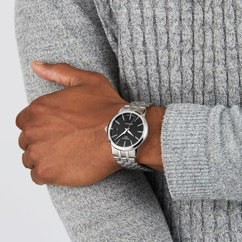 A person wearing a Tissot Classic Dream Swissmatic watch with a silver metal band and black face, dressed in a textured gray sweater, with one arm crossed over their body.