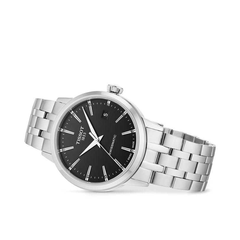 The Tissot Classic Dream Swissmatic watch features a silver stainless steel link bracelet, black dial, and date at 3 o’clock, elegantly displayed at an angle on a white background.