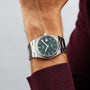 A Tissot PRX Powermatic 80 watch with a green dial and stainless steel case is worn on the left wrist, paired with a burgundy long-sleeve shirt.