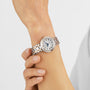 A person adjusts a Tissot Bellissima Automatic watch with a round silver and gold case, white dial, and Roman numerals on their left wrist, highlighting luxury and style against a plain background.