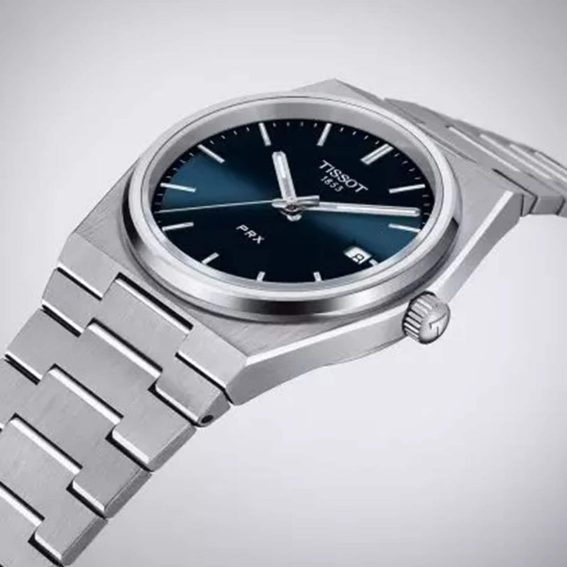 The Tissot PRX is a stainless steel wristwatch featuring a blue dial, silver hour markers, Swiss quartz movement, a date window at 3 o’clock, and a metal link bracelet.