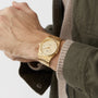 A person wearing the Tissot PRX Powermatic 80 watch with a gold finish and a green jacket over a beige sweater, their hands clasped together.