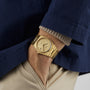 A person in a navy jacket, beige pants, and a gold Tissot PRX Powermatic 80 watch with a textured face and Nivachron hairspring has their hand in their pocket.