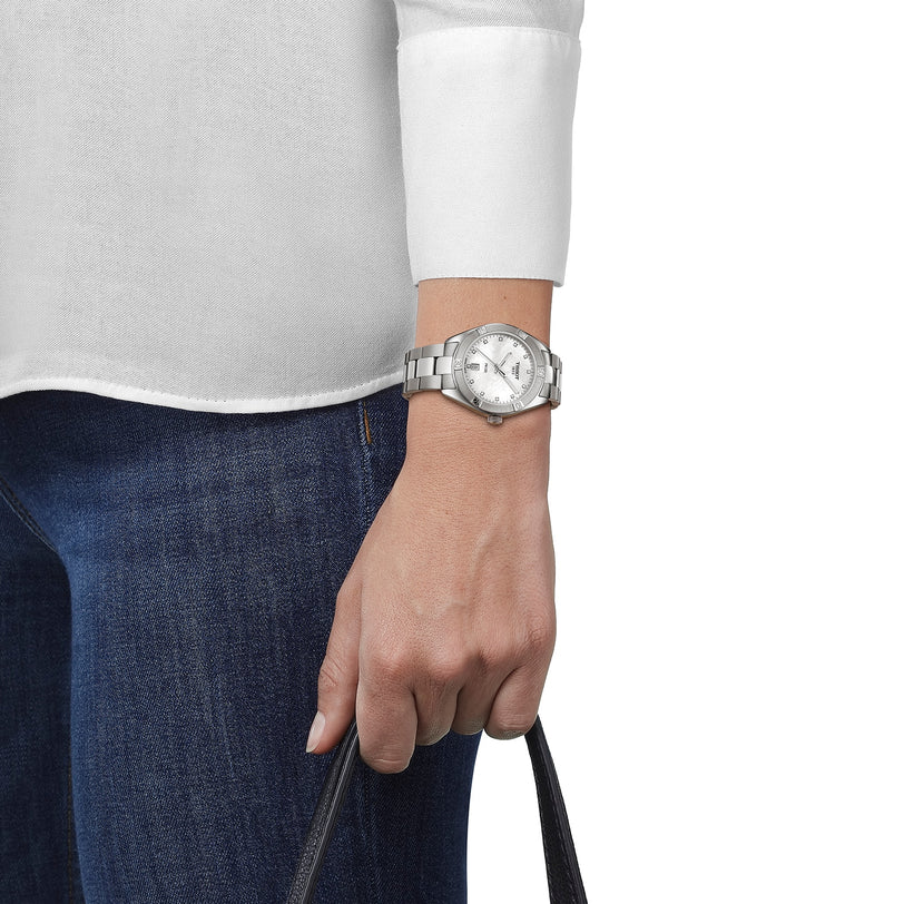 A person in a white shirt and blue jeans holds a black strap, wearing a Tissot PR 100 Sport Chic watch on their left wrist.