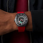 A person in a black jacket showcases a Tissot Sideral S Powermatic 80 watch with a round dial and red strap on their wrist.