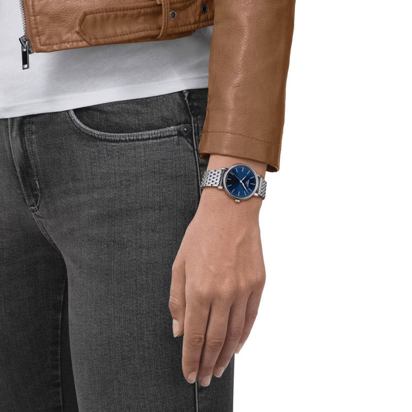 Wearing a brown leather jacket and black jeans, a person showcases the Tissot Everytime—a classic watch with a blue face—on their left wrist.