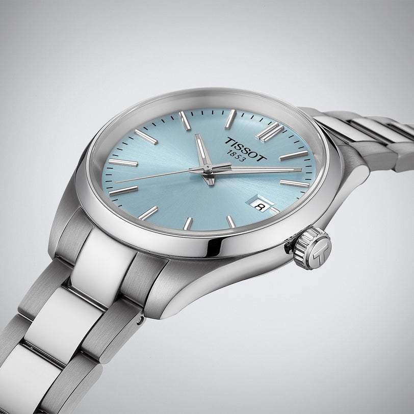 The Tissot PR 100 features a stainless steel case, light blue dial, silver hour markers, date display, and metal bracelet. Powered by Swiss Quartz movement for precise timekeeping.