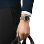 A person in a dark sweater and pants holds a black bag; on their left wrist is a Tissot PR516 Chronograph with a silver and gold finish, black dial, and quartz movement.