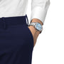 A person in a white shirt and navy pants has their hand in their pocket, showcasing a Tissot PR 100 Chronograph silver wristwatch with a blue face.