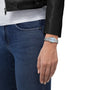 A person stands with hand by their side, wearing blue jeans, a black leather jacket, and a Tissot PRX silver wristwatch.