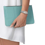 A person wearing a Tissot PRX 25mm silver wristwatch with sapphire crystal holds a light blue clutch purse, shown against a white outfit and plain background.