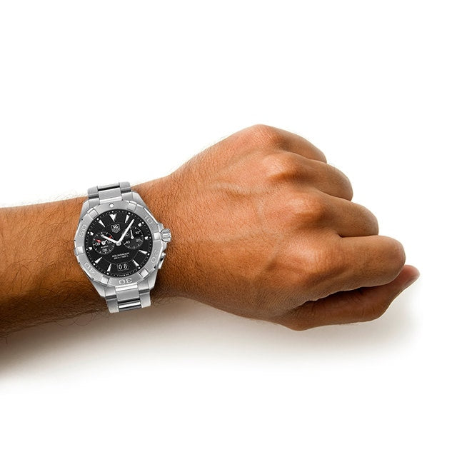 A person’s wrist wearing a TAG Heuer Aquaracer silver metal watch with a black dial, displayed against a white background.