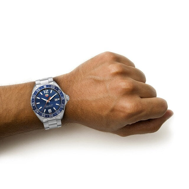 A left wrist wears a silver TAG Heuer Formula 1 Date watch with a blue sunray dial, shown against a white background.