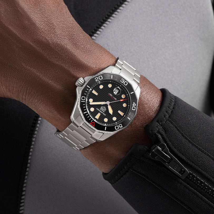 A close-up of a person wearing the TAG Heuer Limited Edition 1 of 500 Aquaracer Professional 300, a luxury tool watch with a silver case and black dial, over a black and grey wetsuit. Features precise calibre 5 automatic movement.