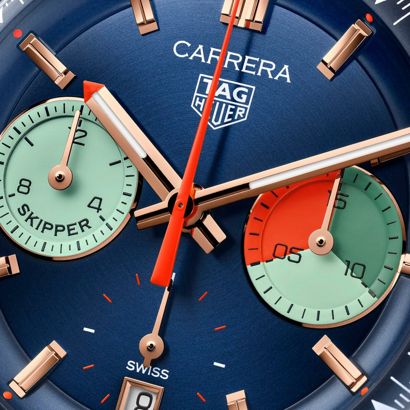Close-up of the TAG Heuer Carrera Chronograph Skipper watch, showcasing gold hands, red accents, and two green SKIPPER and SWISS subdials on a blue face—reflecting its sailing heritage and precision chronograph movement.