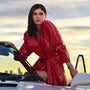 A woman in a shiny red coat leans on a car door at sunset, city skyline blurred behind her, showcasing the TAG Heuer Carrera Chronograph on her wrist.