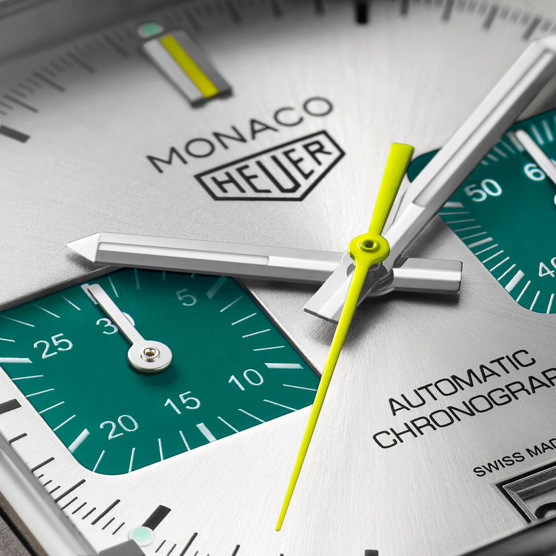 Close-up of a TAG Heuer Monaco Chronograph Racing Green watch face with a silver dial, teal subdials, yellow second hand, and Calibre 11 text plus Monaco, Heuer, and Automatic Chronograph branding.