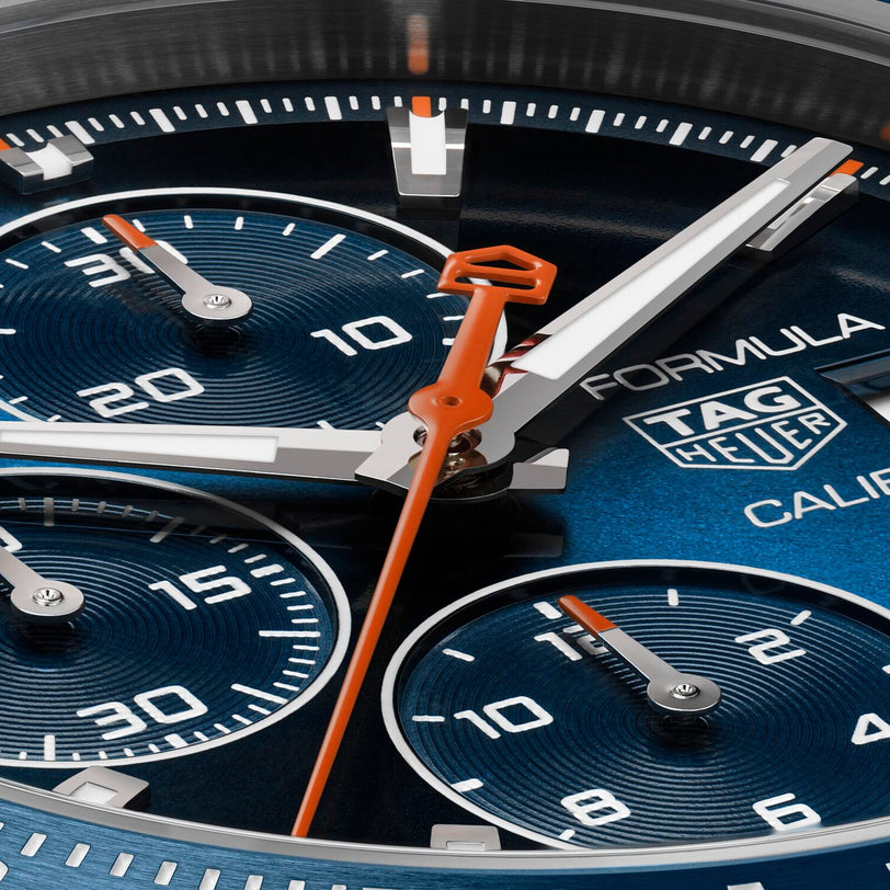Close-up of the TAG Heuer Formula 1 Chronograph watch face, featuring a blue ceramic tachymeter bezel, silver subdials, vivid orange hands, and precise Calibre 16 movement.