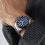 A person wearing a TAG Heuer Formula 1 Chronograph wristwatch with a blue dial and tachymeter bezel, paired with a dark gray knitted sweater.