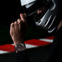 A person in a white racing helmet adjusts their visor, wearing a TAG Heuer Formula 1 Chronograph, as a red and white racetrack barrier in the background underscores the motorsport heritage.