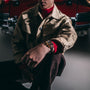 Wearing a beige jacket, a person showcases the TAG Heuer Carrera Chronosprint x Porsche Rallye wristwatch while seated in front of a red vintage car.