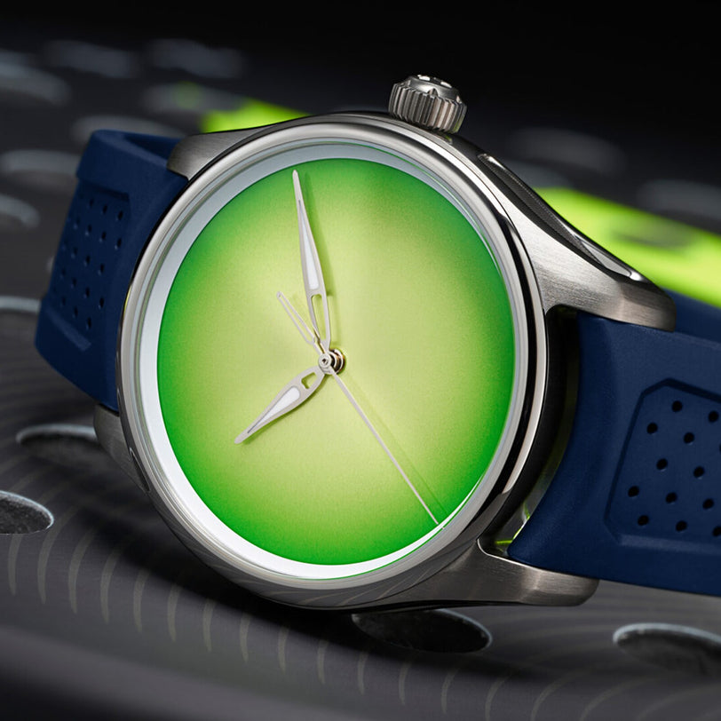 The H. Moser & Cie Pioneer Centre Seconds Concept Citrus Green wristwatch features a green gradient dial and blue strap, displayed on a dark, perforated surface.
