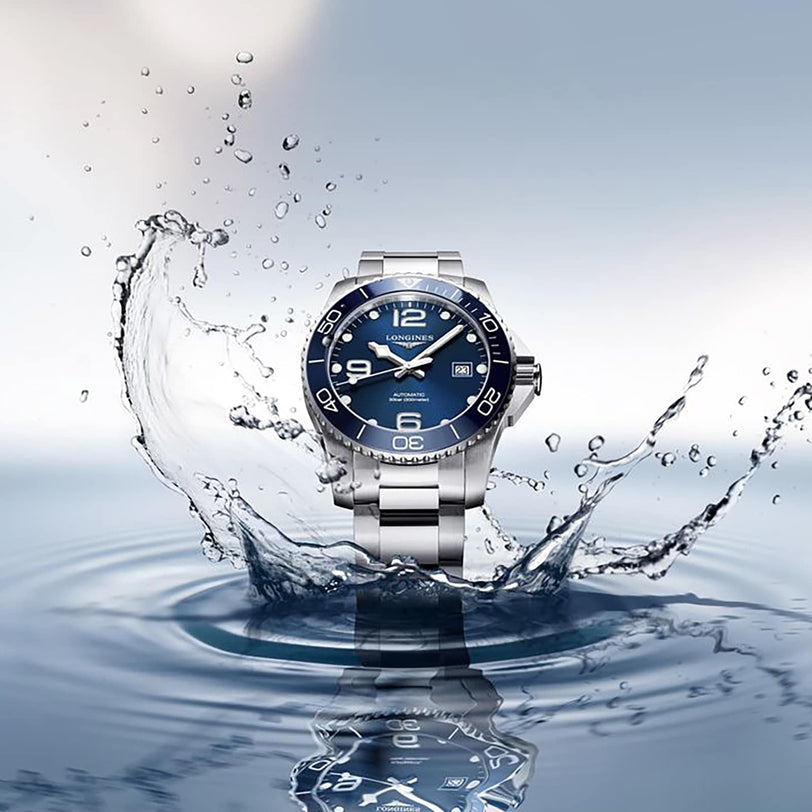 The Longines HydroConquest, a stainless steel diving watch with a blue face, is showcased as water splashes on a reflective surface, emphasizing its water-resistant design.