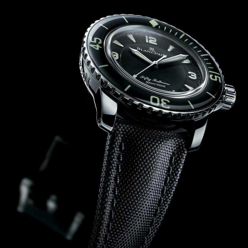 A close-up of the Blancpain Fifty Fathoms Automatique shows its 45mm steel case, black dial with luminous markers, and textured black strap set against a black background.